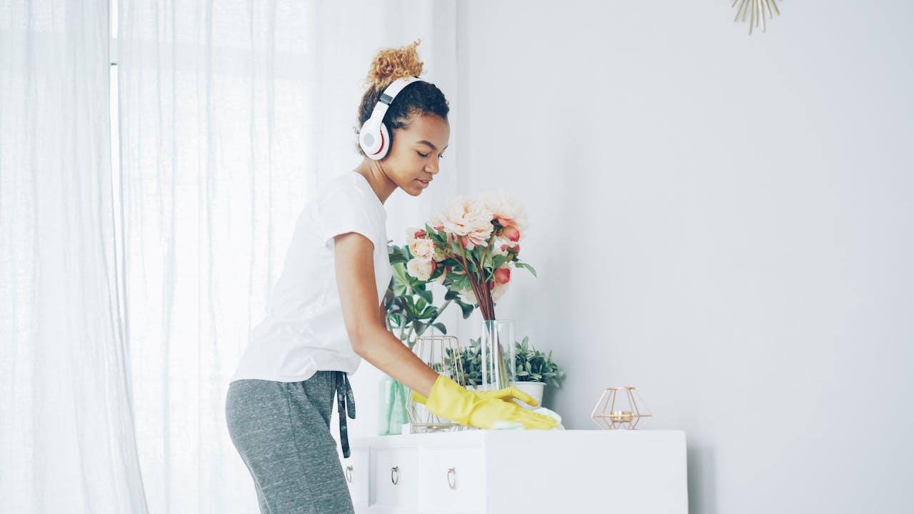 Woman in white shirt cleans table while listening to music with headphones. Bright indoor scene.